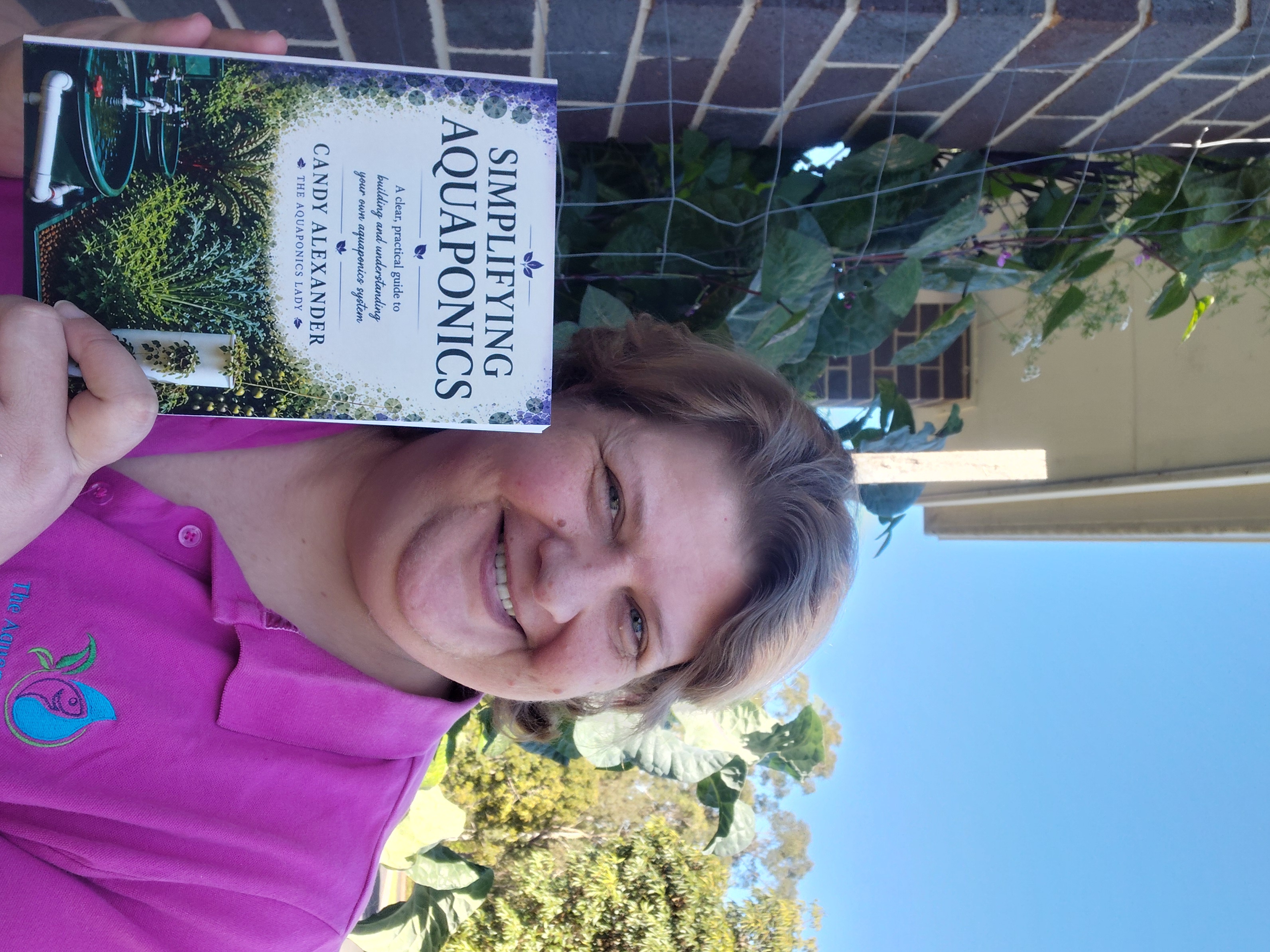 Candy Alexander, The Aquaponics Lady, holding her Simplifying Aquaponics book beside her garden.