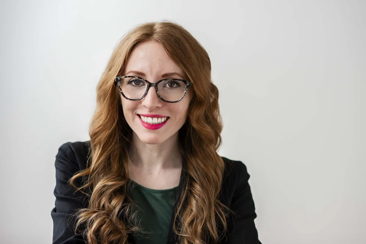 Amber Krasinski - red hair woman with brown glasses and pink lipstick wearing a black blazer and smiling