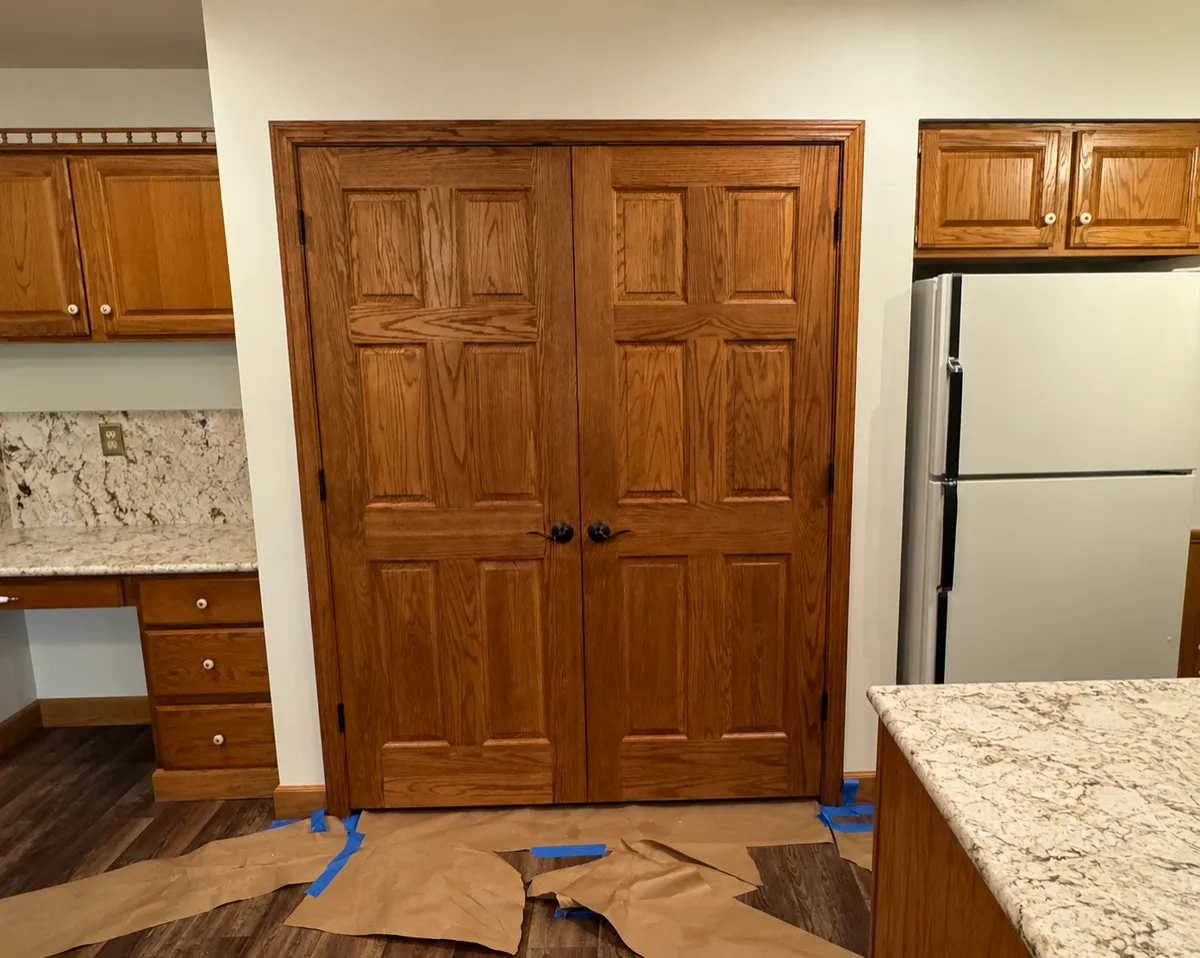 Natural oak double pantry doors with raised wood panels and dark hardware in a residential kitchen, showcasing detailed woodwork and door refinishing.
