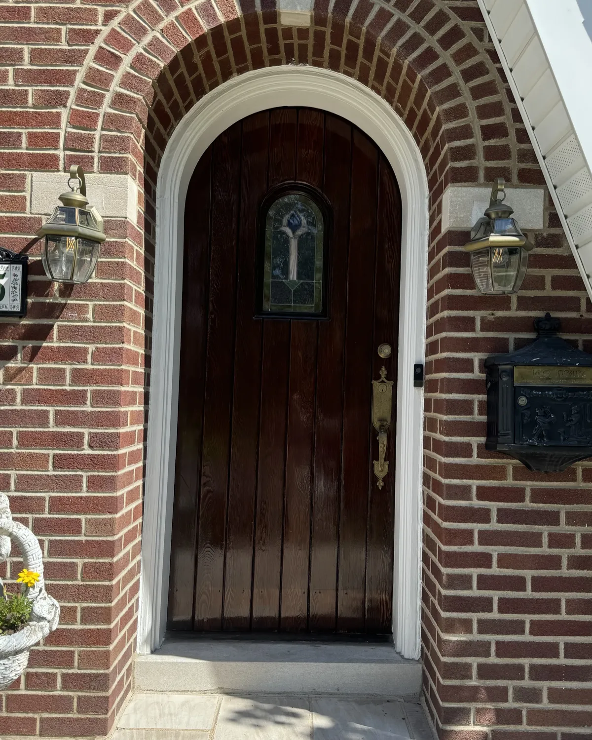Arched solid wood front entry door with rich dark brown stain, decorative glass inset, and white trim surrounded by red brick.