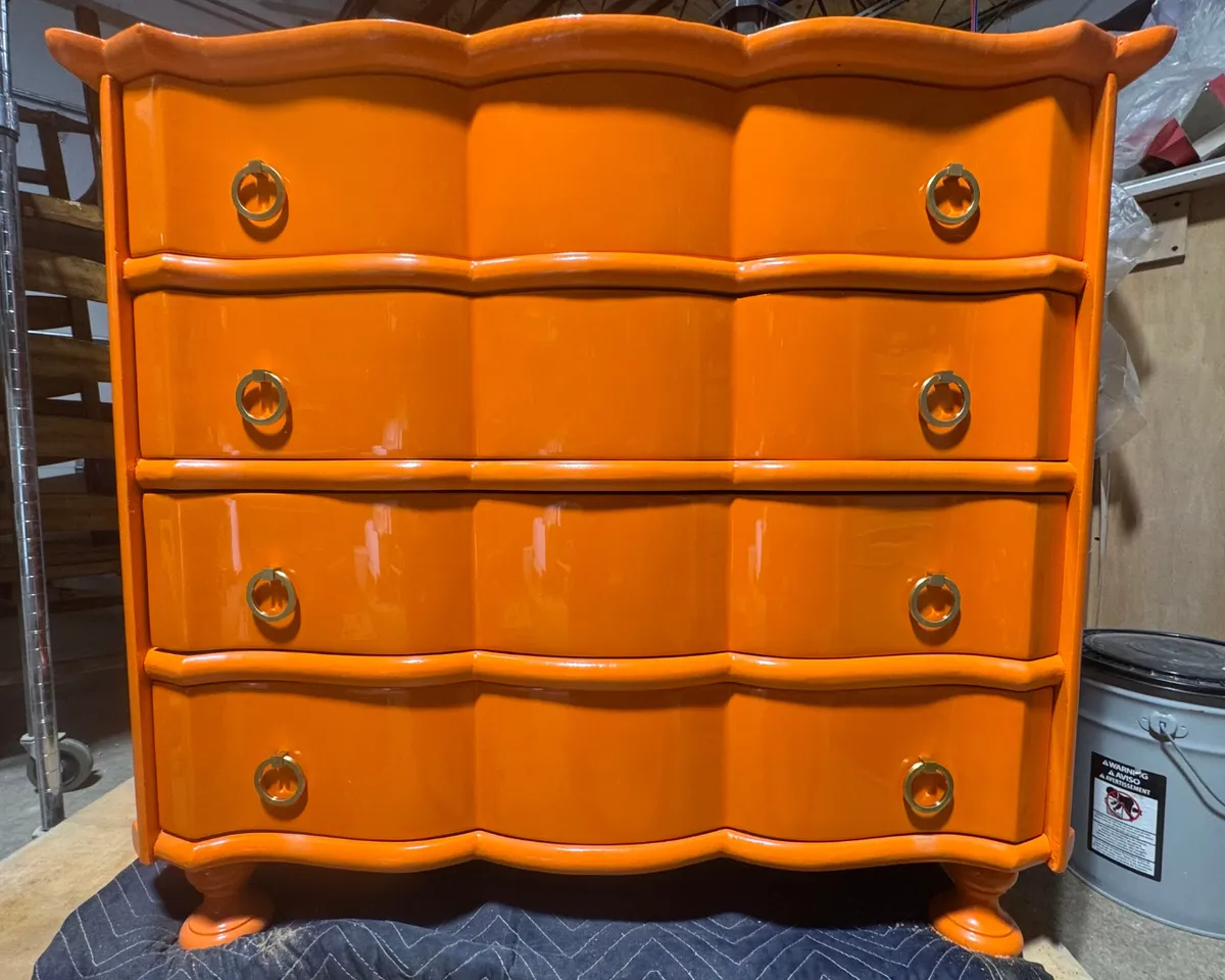 Bright orange refinished vintage wooden dresser with curved drawer fronts and decorative metal hardware, showcasing custom furniture painting and refinishing.