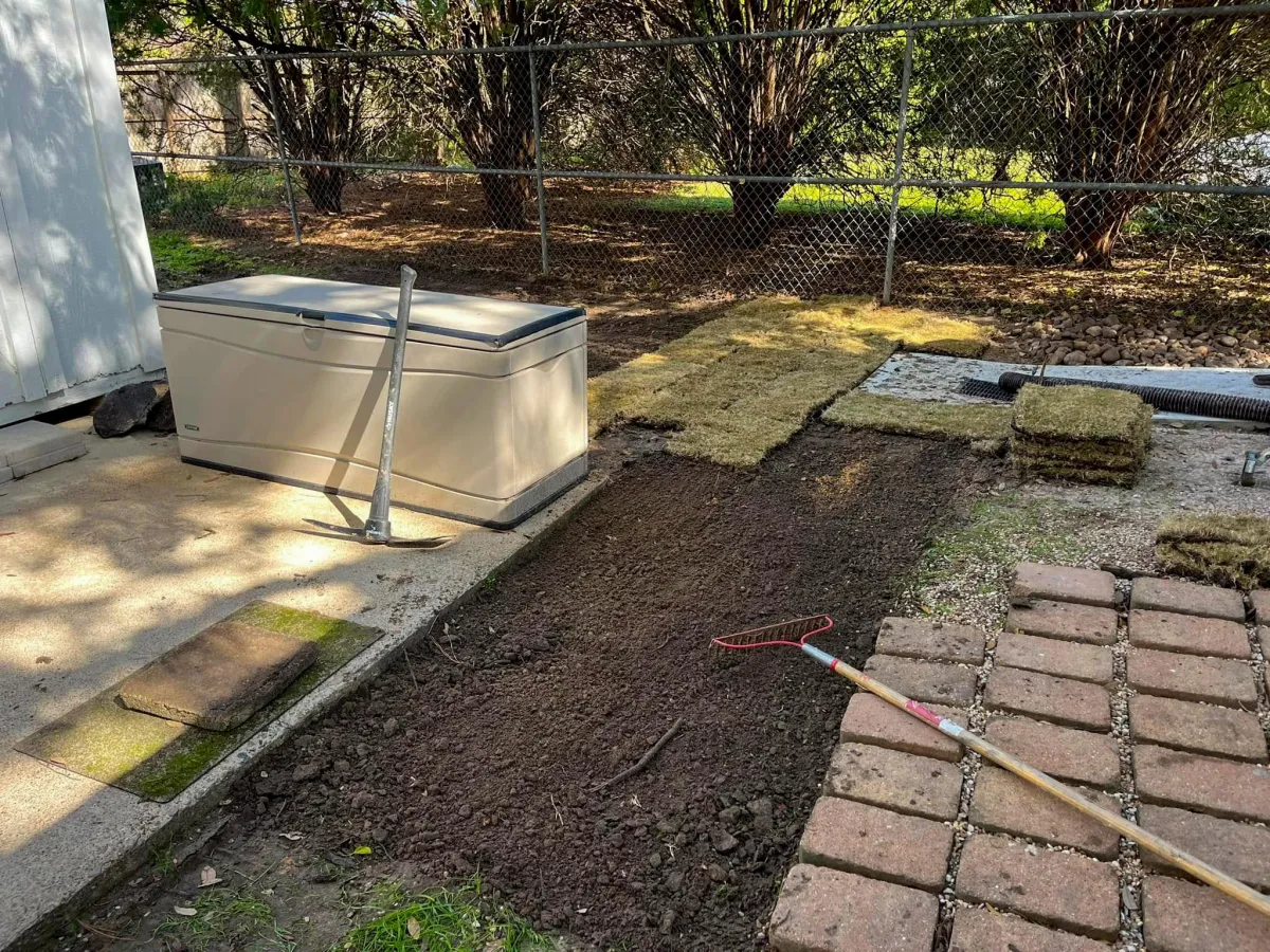 Landscaping in progress—freshly tilled soil strip between patio and brick pavers, with stacked sod pieces, rake, and pickaxe ready for installation in a Houston backyard.