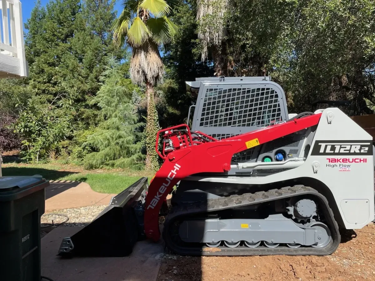 Skid steers excavation in a backyard surrounded by trees.