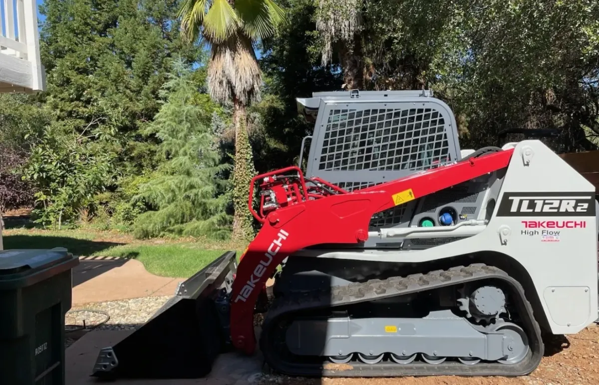 Skid steers excavation in a backyard surrounded by trees.