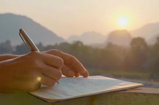 A person writing in a journal with a pen at sunrise, overlooking a peaceful mountain landscape.