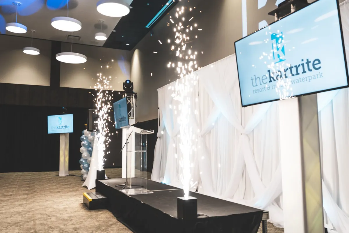 Cold spark fountains on a corporate event stage beside a podium and branded screens at The Kartrite