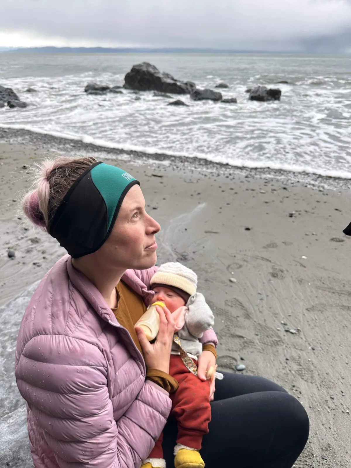 A person feeding a baby with a bottle on a beach with grey sand and grey sky. They are wearing black leggings and a pink puffer jacket.
