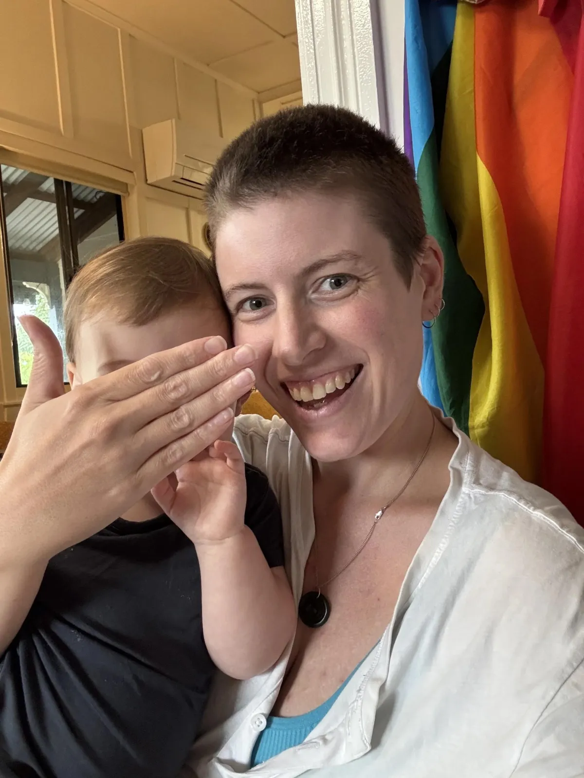 A person with a shaved head smiling with teeth, with a child on their lap. The person's hand is covering the baby's face. There is a rainbow pride flag behind them.