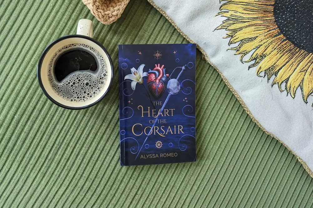 a book titled "The Heart of the Corsair" by Alyssa Romeo sits on a background of green corduroy next to a coffee cup and sunflower pillow