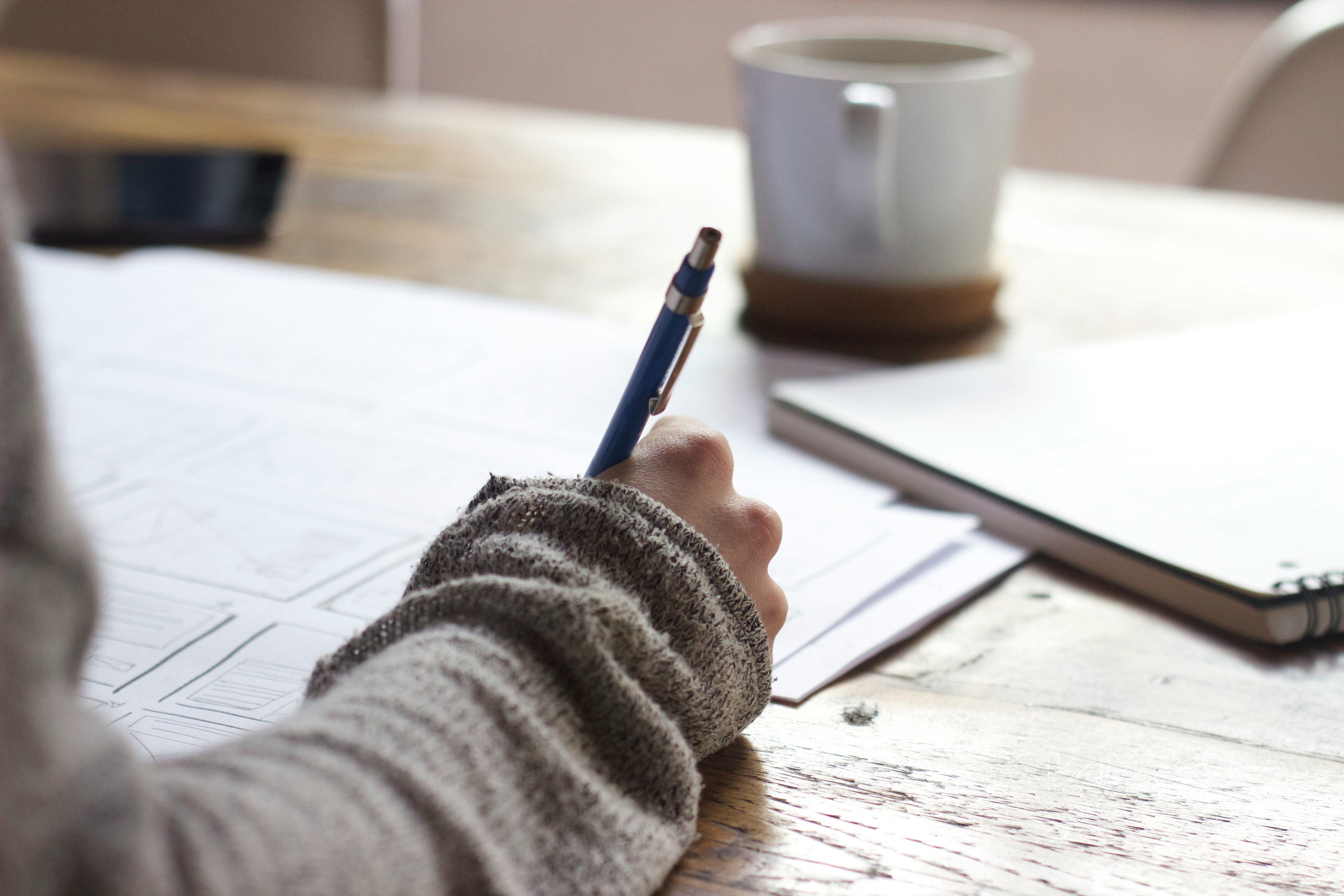 A sweater-clad hand sketching out ideas on paper, coffee cooling nearby