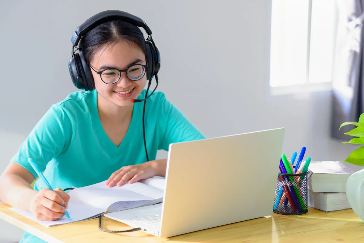 A girl attending an online class smiling and taking notes