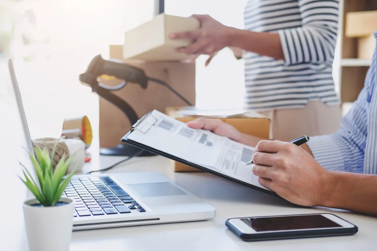 A guy analysing the statistic sitting at the table where a laptop is placed and a lady standing beside him carrying a box