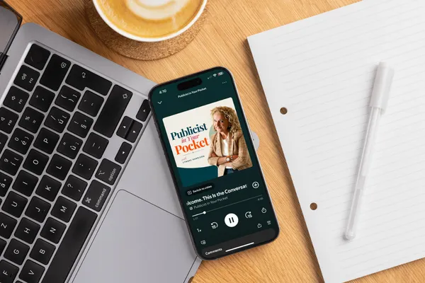 Smartphone displaying the Publicist in Your Pocket podcast beside a laptop, notebook, and coffee on a desk workspace