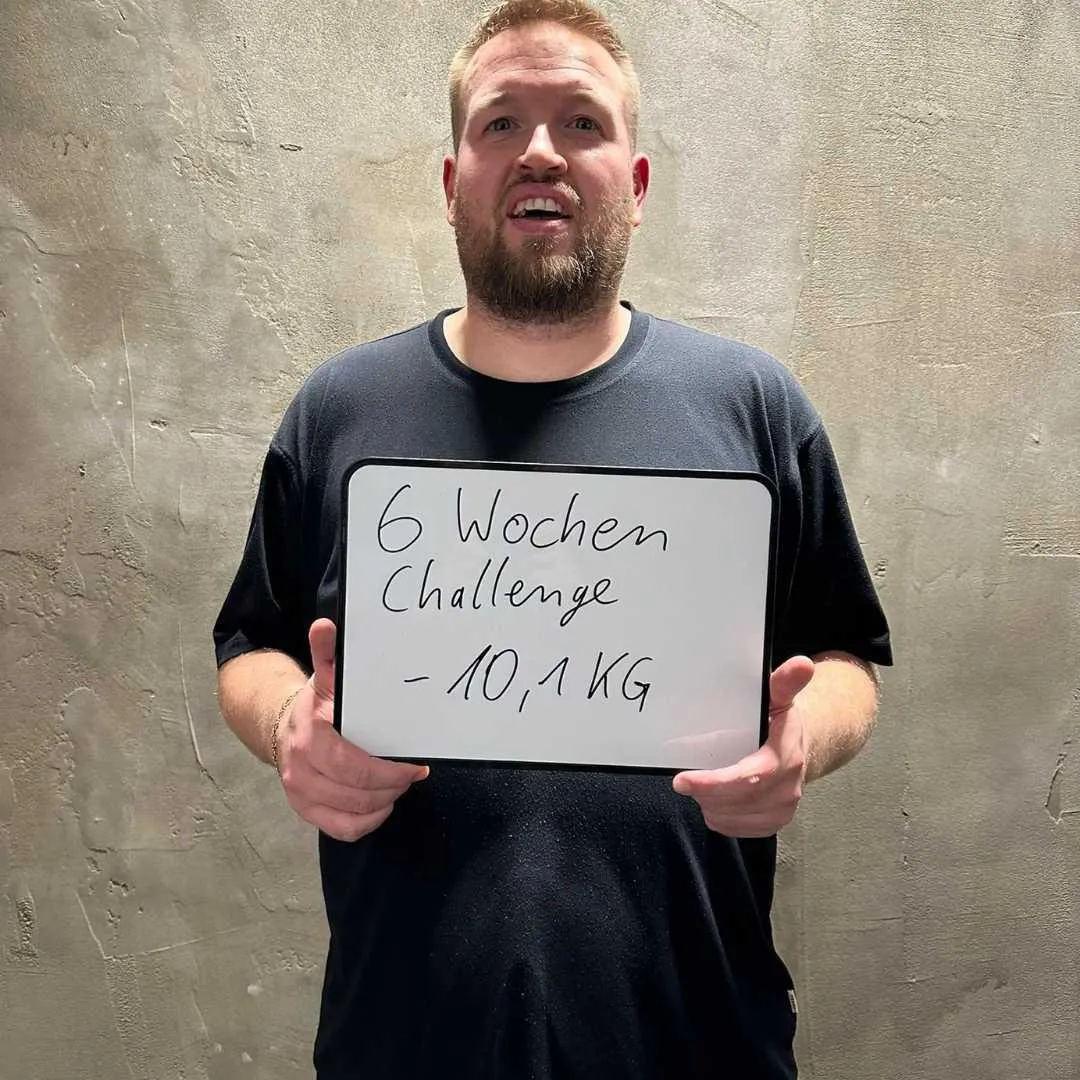 Ein Mann in einem schwarzen T-Shirt hält ein Schild mit der Aufschrift „6 Wochen Challenge - 10,1 KG“. Er zeigt eine stolze Haltung vor einer grauen Betonwand und feiert seinen beeindruckenden Gewichtsverlust.