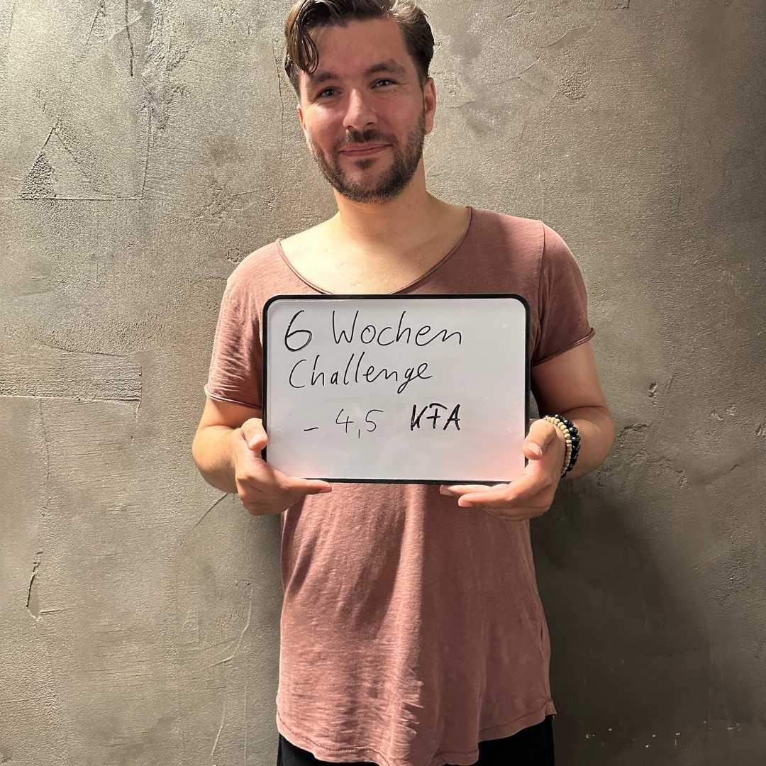 Ein Mann in einem braunen T-Shirt hält ein Schild mit der Aufschrift „6 Wochen Challenge - 4,5% KFA“. Er lächelt leicht, steht vor einer neutralen Betonwand und präsentiert