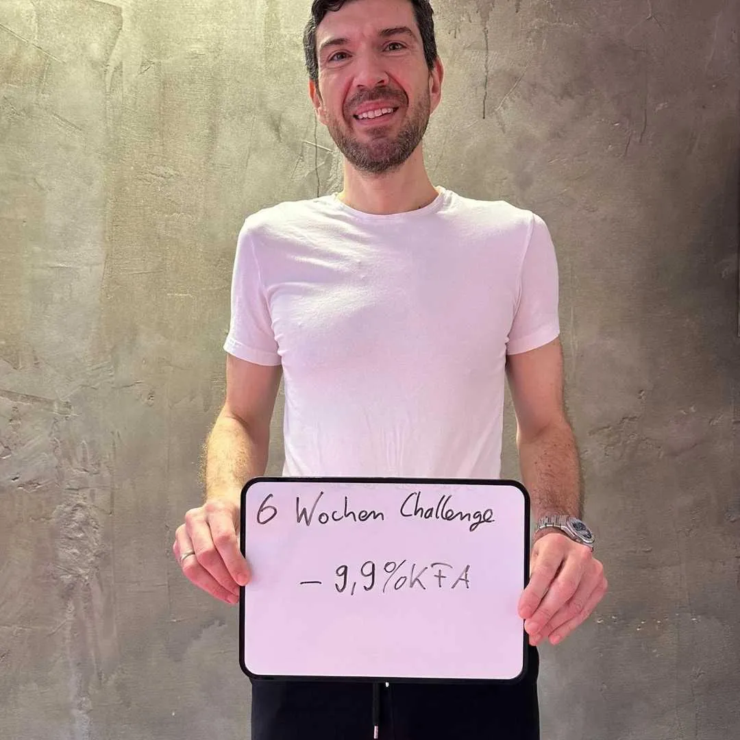 Ein Mann in einem weißen T-Shirt hält ein Schild mit der Aufschrift „6 Wochen Challenge - 9,9% KFA“. Mit einem zufriedenen Lächeln und vor einer Betonwand präsentiert er den Erfolg seiner Fitness-Transformation.