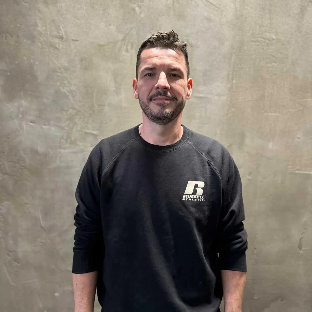 Ein Mann in einem schwarzen Sweatshirt steht vor einer grauen Betonwand. Mit einer entspannten Haltung repräsentiert er das Ergebnis kontinuierlicher Fitnessbemühungen.