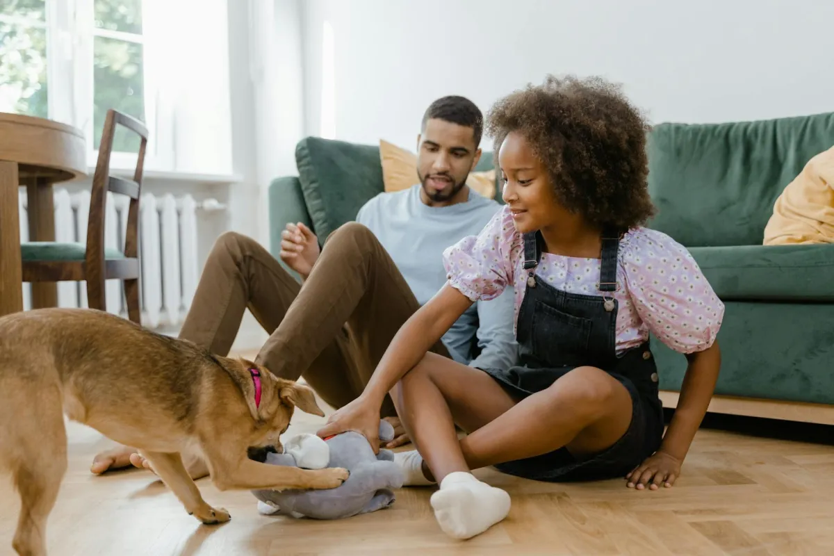 A girl and man playing with a dog on the floor in a white room near a green sofa, table, and chairs.