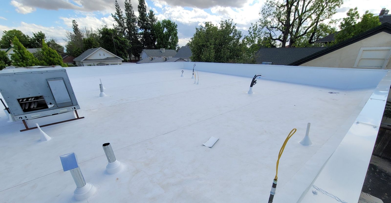 White low-slope roof with sealed pipe vents and a rooftop unit, with trees and houses behind