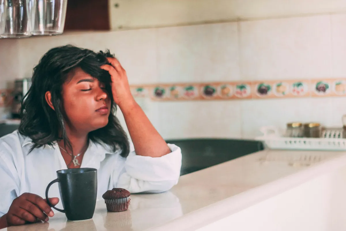 Woman sitting at counter with coffee mug, muffin, and holding head