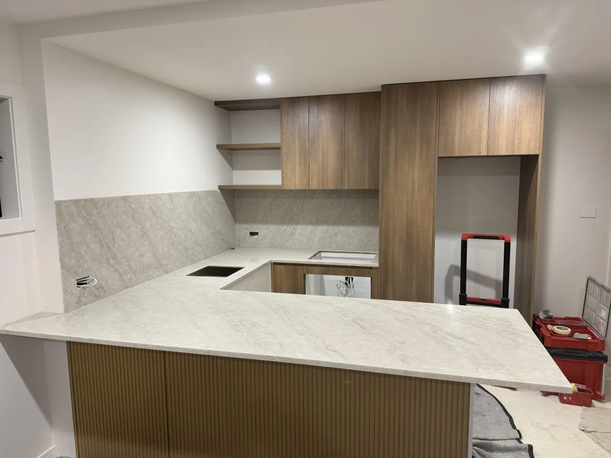 Modern kitchen with polished beige stone countertops and backsplash, warm wood cabinetry, and minimalist lighting, showcasing the Desert Bloom Counter Surface with soft golden undertones.