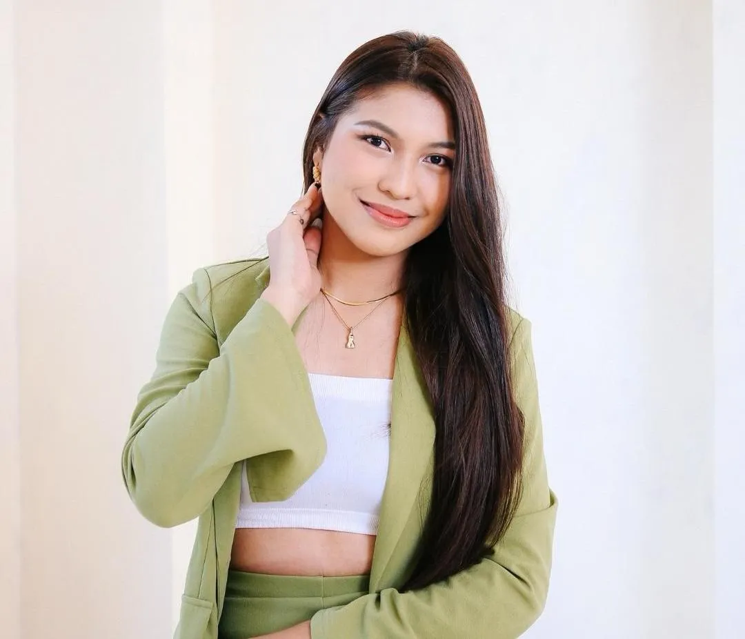 A young woman with long, straight dark hair smiles softly at the camera. She is wearing a light green blazer over a white crop top, accessorised with gold jewellery, and stands against a softly lit, neutral background.
