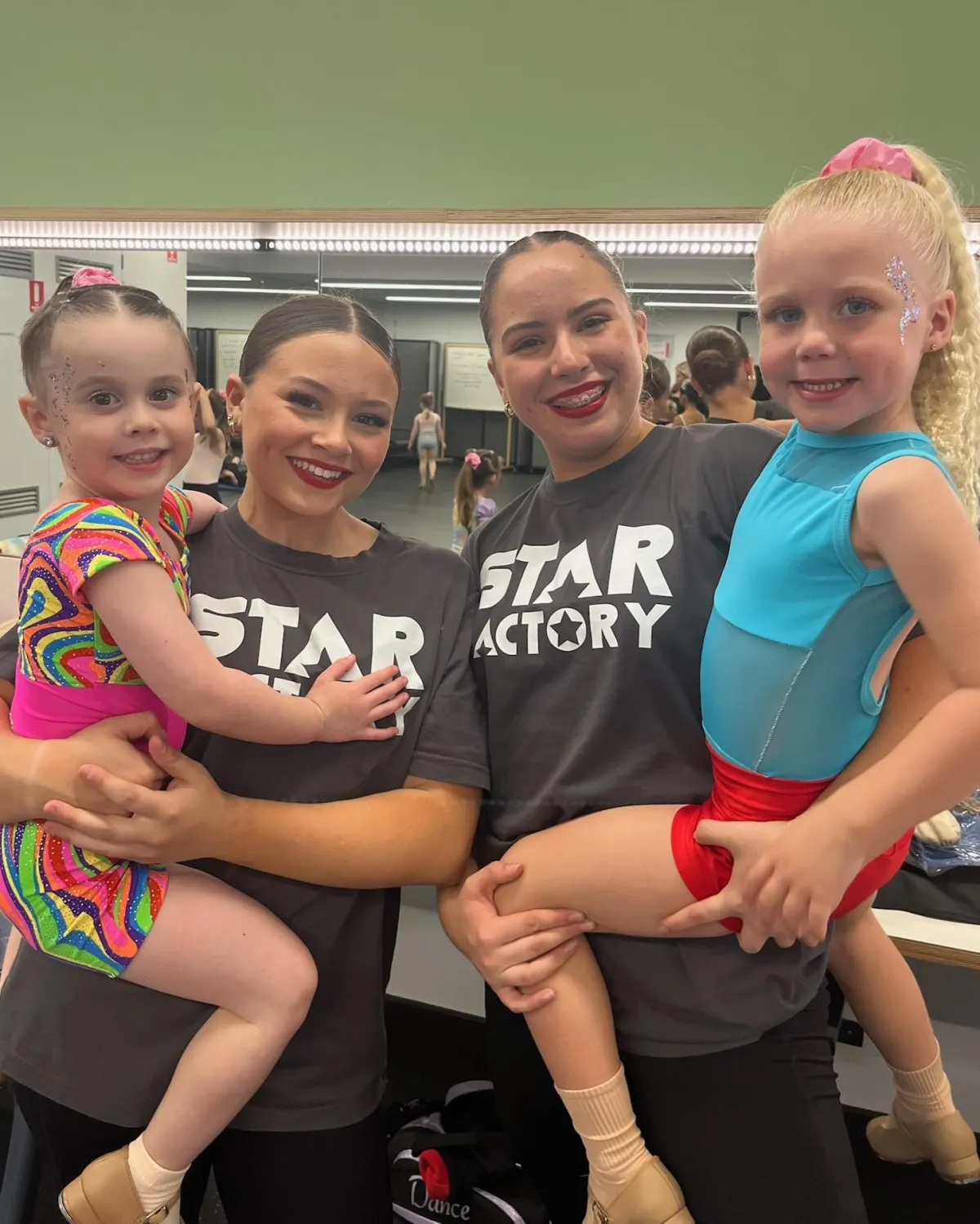 Two Star Factory instructors holding young dancers in bright, glittery face make-ups, posing backstage with big smiles. The vibrant makeup and colourful outfits highlight the excitement of performance day, reflecting The Star Factory’s commitment to building confidence and stage presence in their students through dance and cheer.