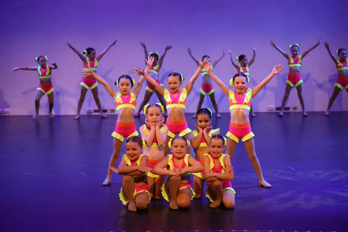 Smiling young dancers in vibrant pink and yellow costumes light up the stage during a jazz performance, showcasing the fun, energy, and stage presence that define our recreational jazz dance classes.