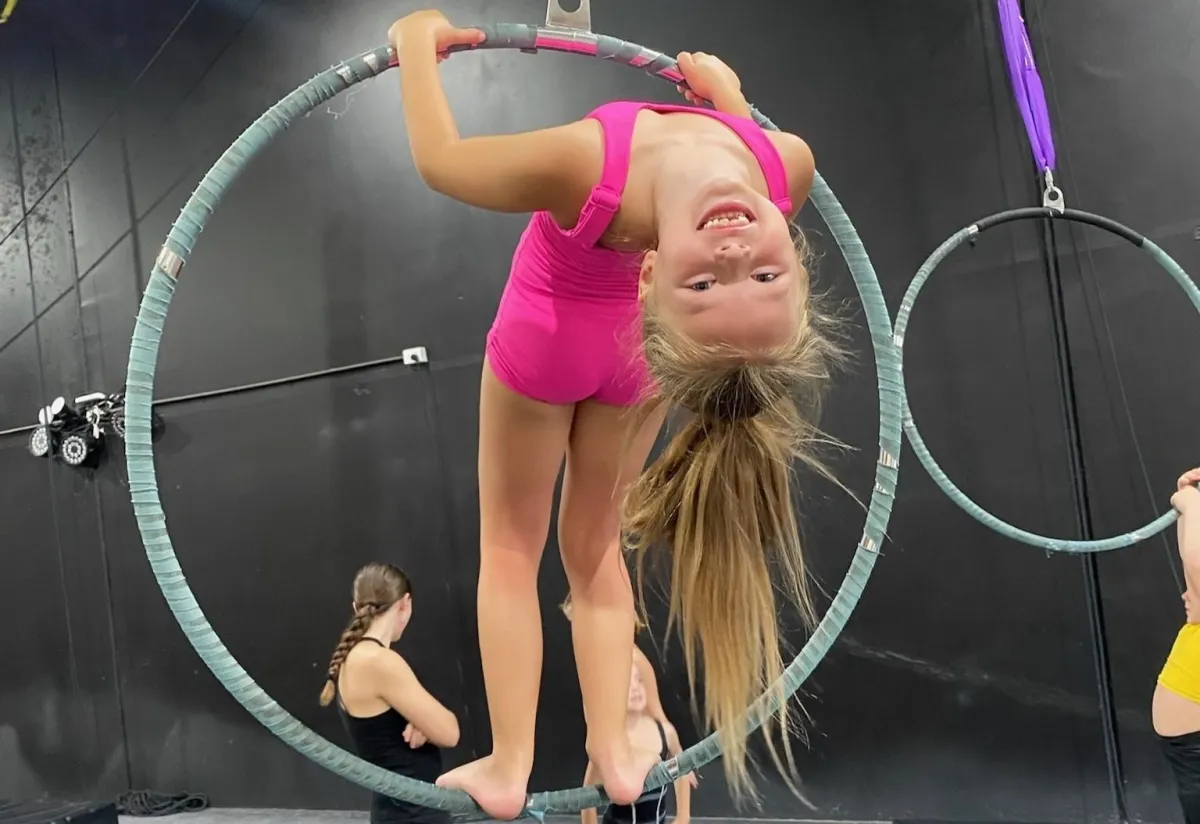 A young girl smiling while hanging upside down in a hoop during aerials class at The Star Factory Joondalup, capturing the magic, strength, and thrill of recreational aerial silks and hoop training.