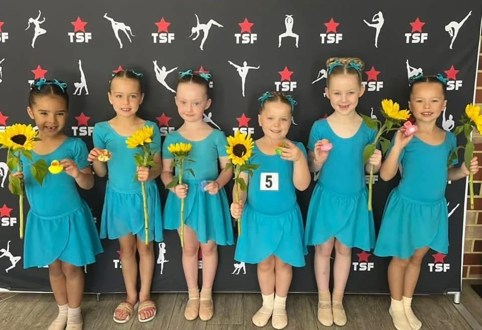 Smiling ballet students in blue uniforms holding sunflowers and plush toys after completing their dance exams, marking a joyful milestone in Comdance and RAD training at The Star Factory Joondalup.
