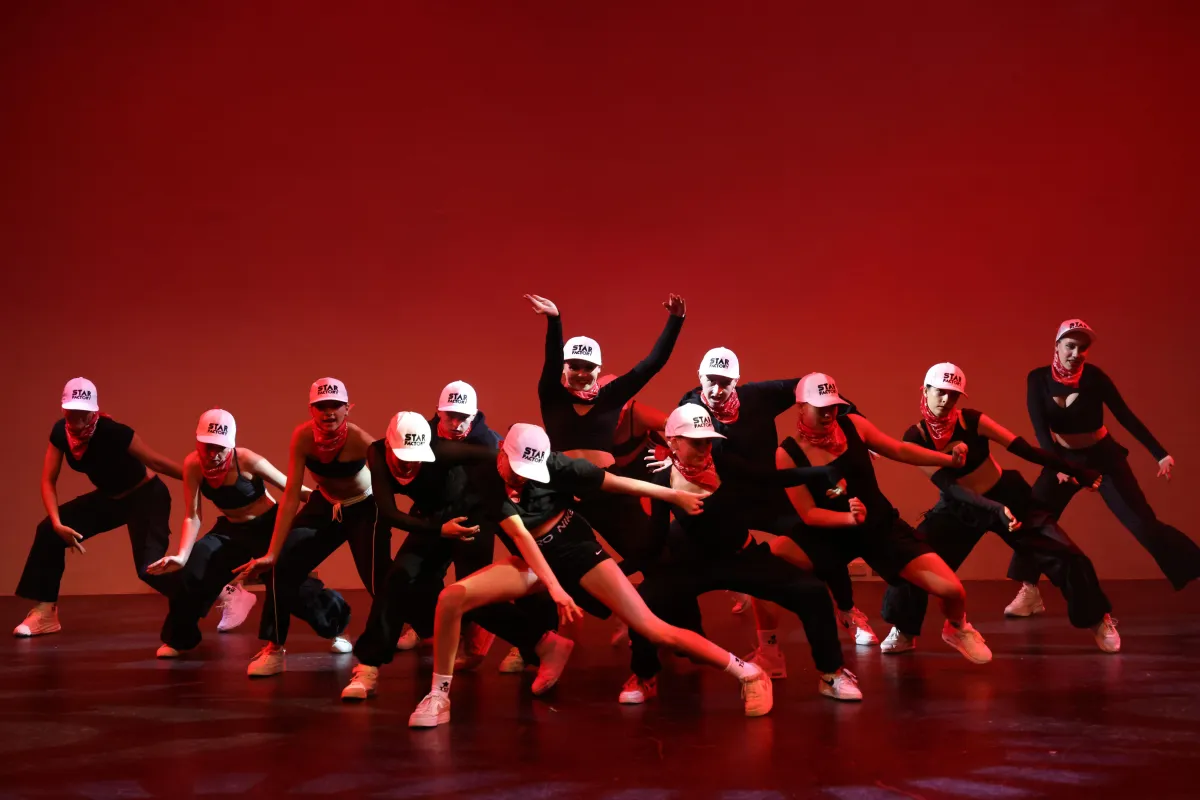 A hip hop crew of teen and young adult dancers perform a powerful V-formation routine in black outfits and white caps on stage, embodying boldness, unity, and individuality through urban dance at The Star Factory Joondalup.
