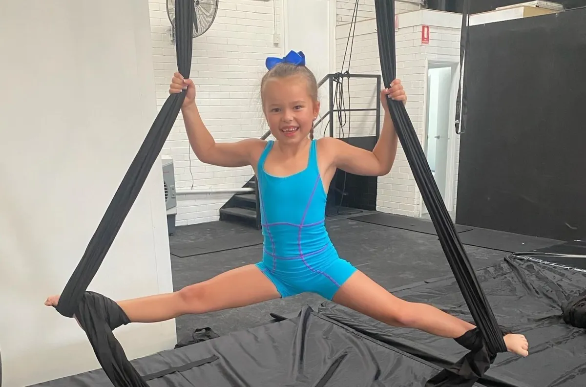 A smiling young girl in blue uniform hangs from aerial silks wrapped around her feet during class at The Star Factory Joondalup, showcasing grace, confidence, and joy in our aerial dance program.