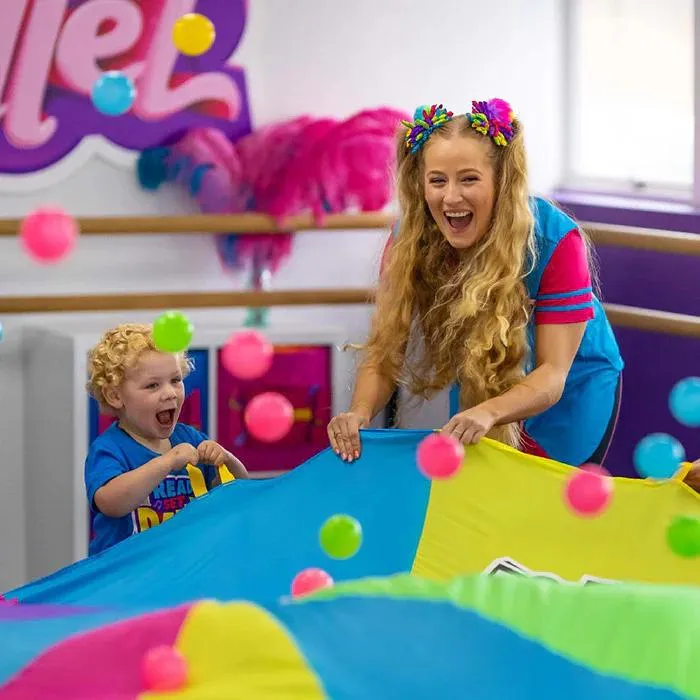 A woman and a young boy bounce colourful balls together using a bright multi-colored cloth. A joyful burst of teamwork and giggles, where simple play builds connection and early motor skills.