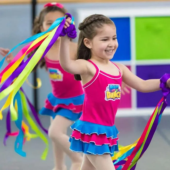 A young girl in a bright pink and blue dance outfit smiles joyfully while waving colourful rainbow ribbons during class, capturing the pure delight and boundless energy of early dance, where every ribbon wave feels like magic in motion.