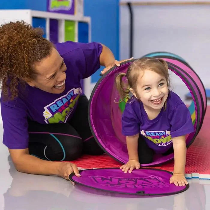 A smiling instructor kneels beside a young girl crawling through a purple play tunnel during an activity. A warm playful moment showing the trust and joy between teacher and child, where learning happens through laughter.
