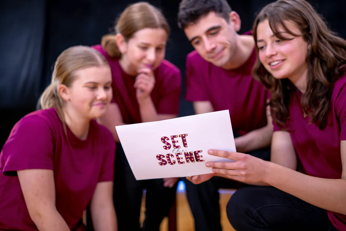 Four young adults in purple shirts huddled curiously around a Set The Scene card, sharing intrigue and anticipation during the acting workshop.