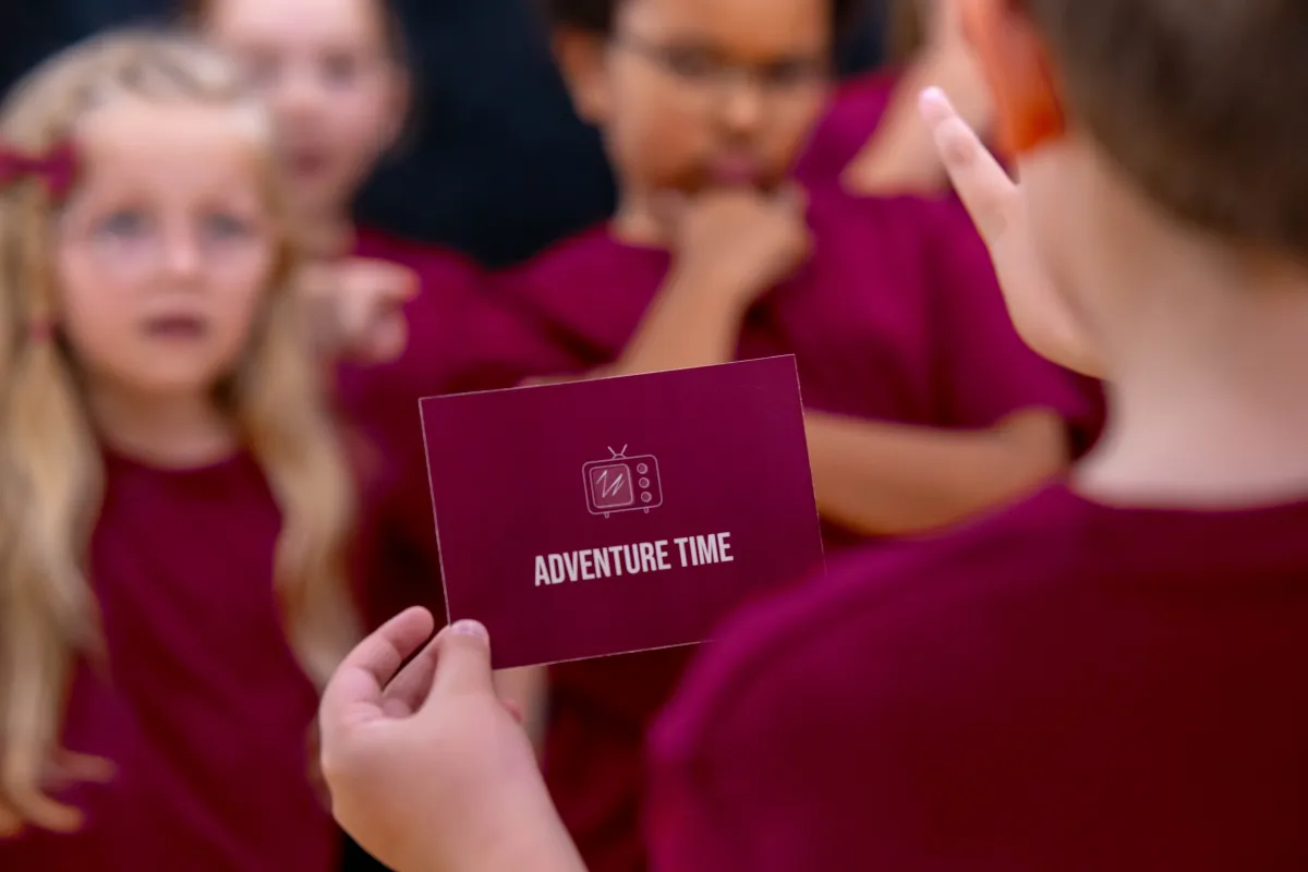 Boy animatedly acting out “Adventure Time” in a playful charades game, with children laughing and guessing in the Set The Scene class.