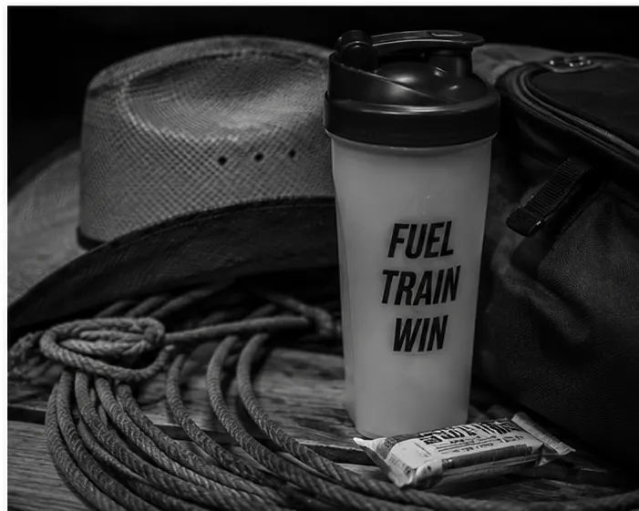 Black-and-white photo of a protein shaker bottle beside a cowboy hat, rope, and gear, representing nutrition and preparation for rodeo athletes.