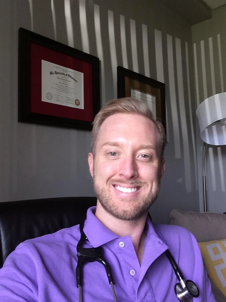Smiling male healthcare professional in a purple polo shirt with a stethoscope, seated in an office with framed diplomas and soft lighting, representing a welcoming atmosphere at Venus Spa.