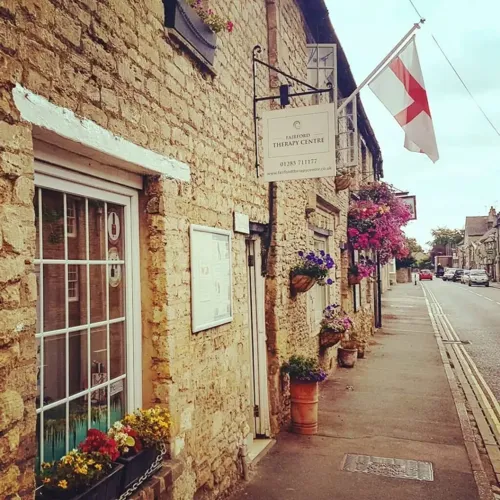 Exterior of Fairford Therapy Centre on a traditional Cotswold stone street with hanging baskets and floral displays, home to The Considerate Practice clinic appointments