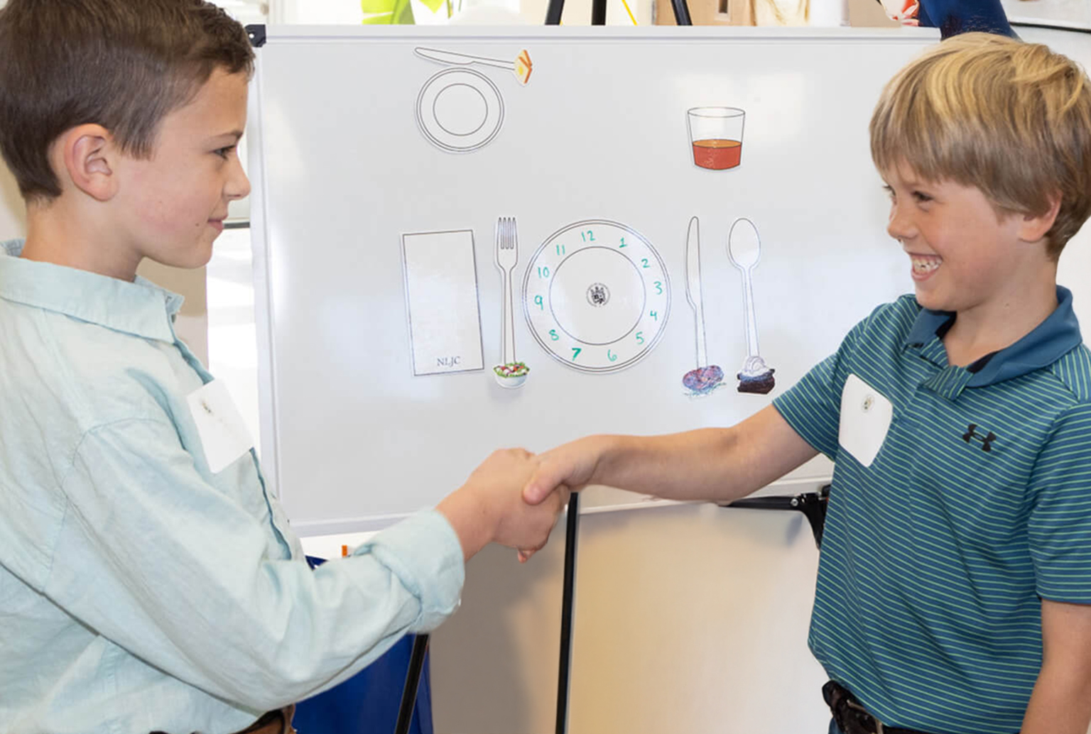Young boys gentlemen shaking hands in front of a table place setting smiling at each other making eye contact Bentwood, TN Pre-Cotillion Manners Etiquette Grades 1-5