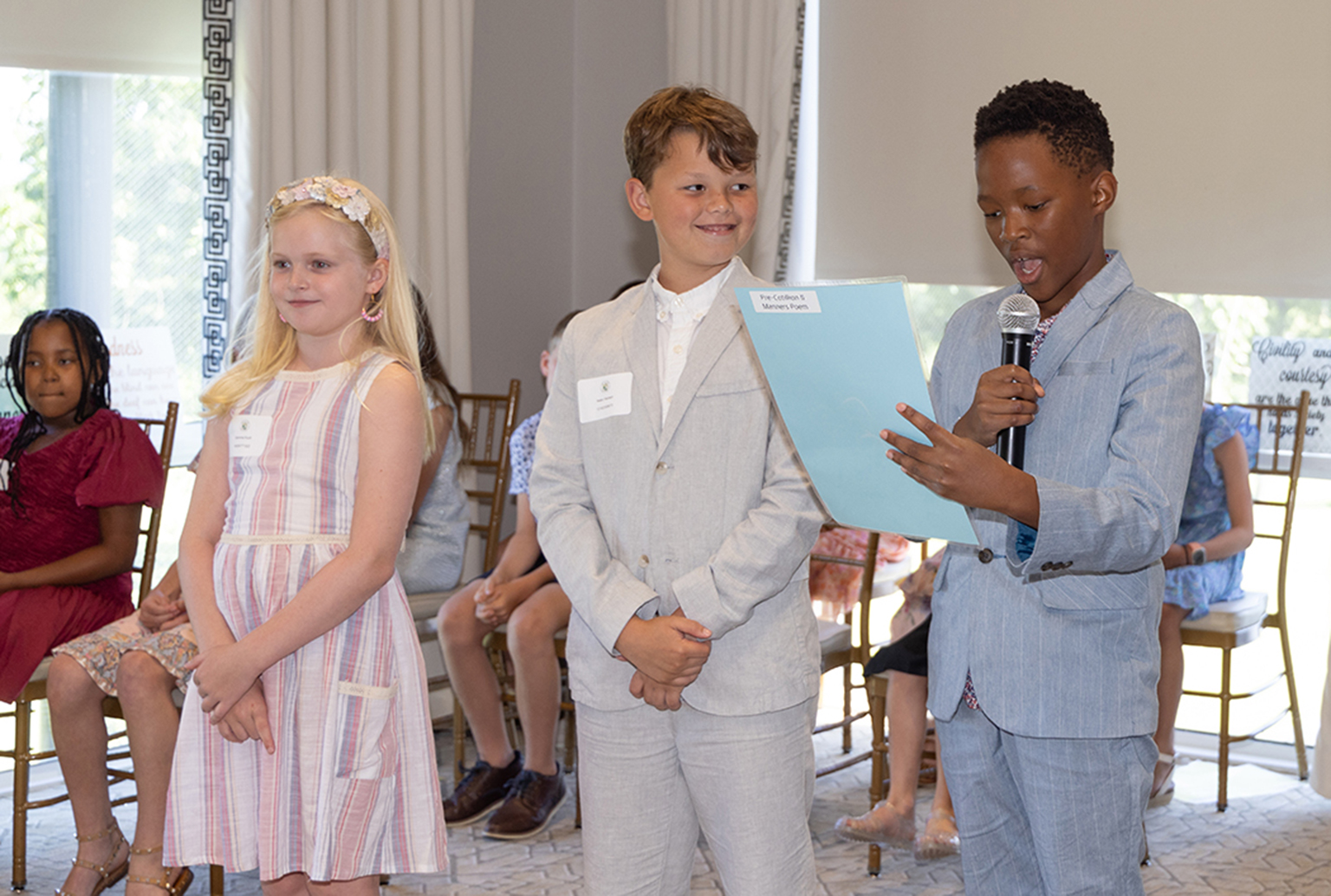 Young gentlemen and ladies speaking in front of a group of parents for Pre-Cotillion NLJC Manners Camp Graduation Day Smiling confidently 