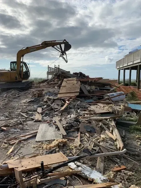 Demolished house ruins in Myrtle Beach with twisted metal debris, coastal background, construction equipment, and lush greenery