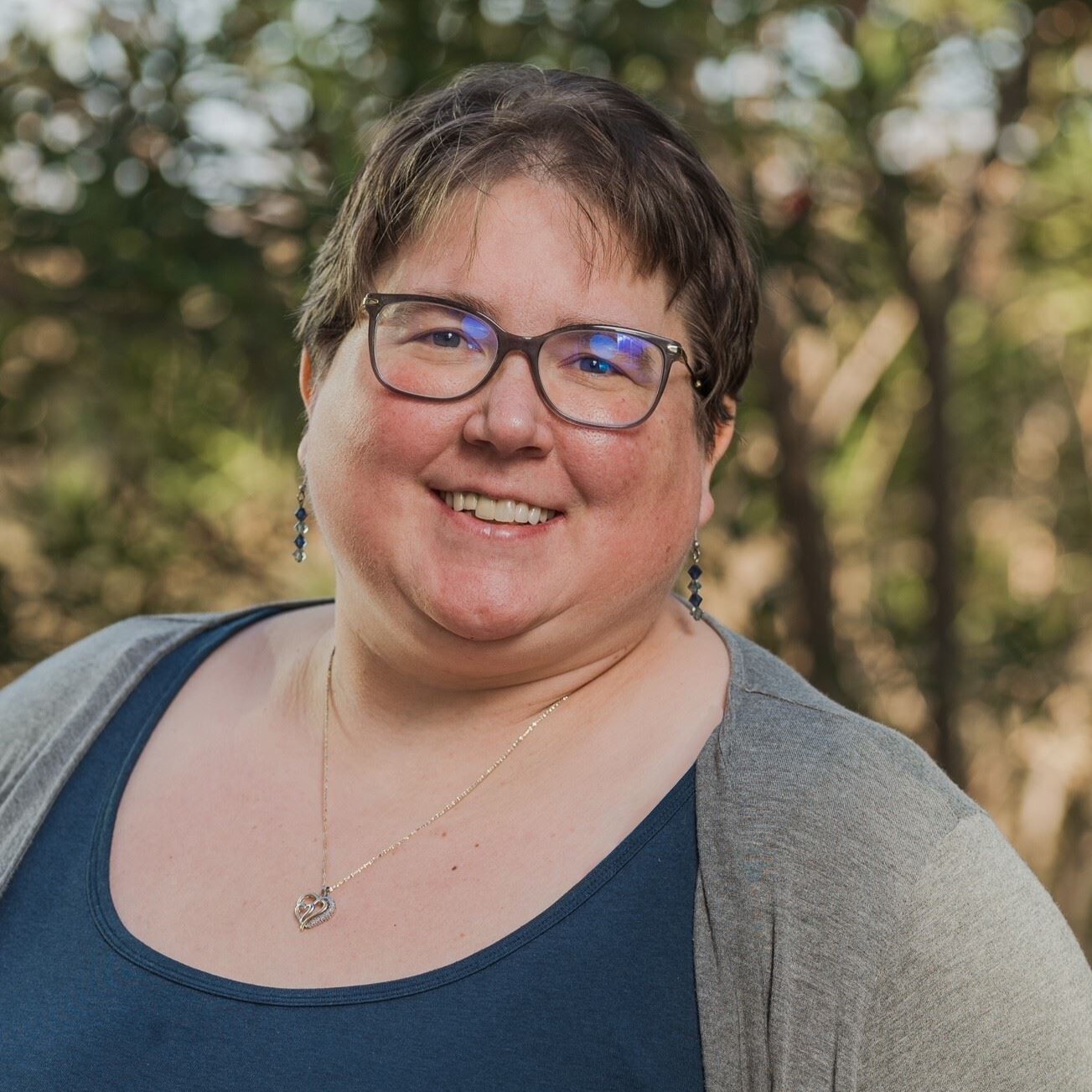 Jenn Davey is a white woman with short brown hair wearing glasses, earrings and a heart pendant necklace.