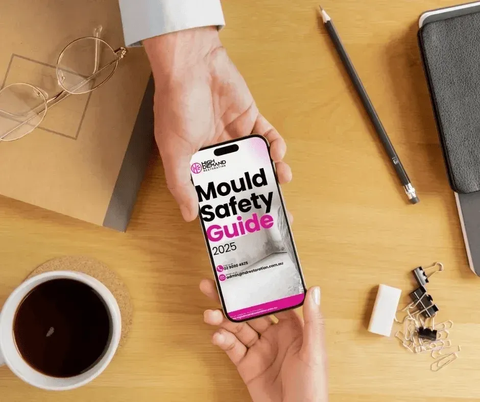 Person holding phone showing High Demand Restoration’s Mould Safety Guide 2025 next to desk and coffee cup.