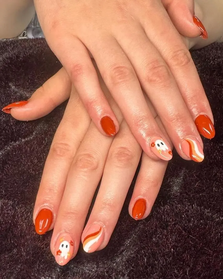 a set of orange fall nails. Three nails are solid orange, one is nude with white and orange stripes, and the final nail has a white ghost and two orange flowers