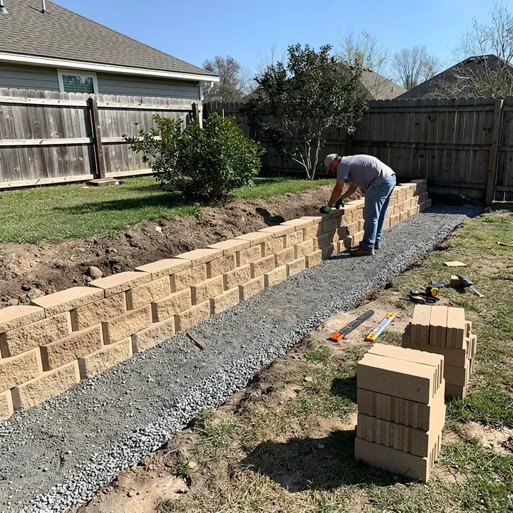 Retaining wall installation in progress along a sloped yard in Denham Springs with worker stacking blocks in the distance