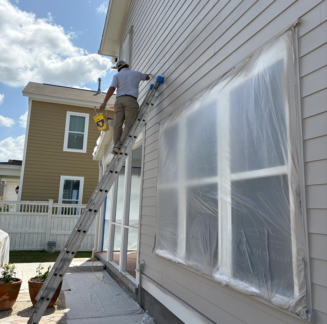 Exterior painter on a ladder repainting house siding with windows protected for prep in Summerville SC