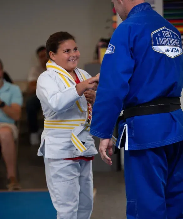 Young student smiling while receiving her belt from a coach at Fort Lauderdale Jiu-Jitsu kids program