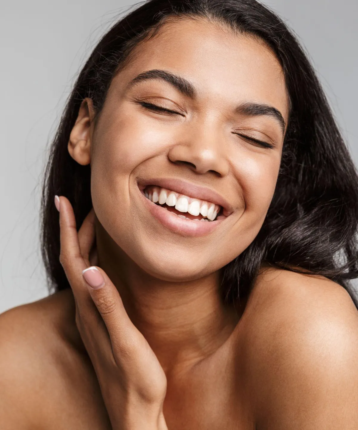 Smiling woman with long dark hair and eyes closed, gently touching her face against a light gray background—evoking serenity and refreshed appearance.